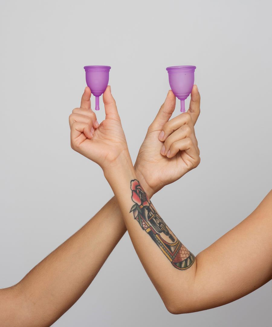 Two people holding a small and large purple Silicone, BPA free and latex free Ruby Cup in between their fingers. The image shows their arms crossing over and is on a light grey background