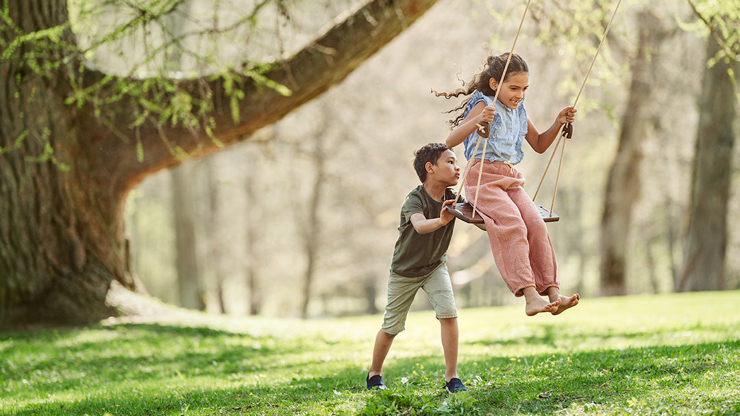 A little boy pushing a little girl on a Lillagunga swing, which is outdoors and attached to a tree