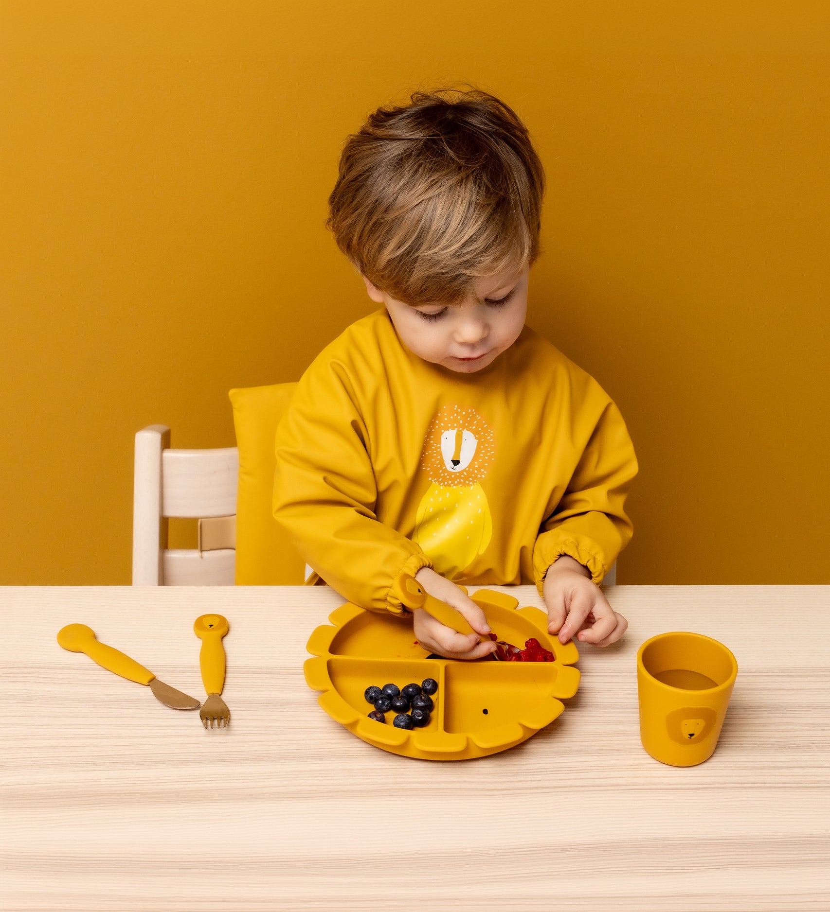 Child wearing Trixie Animal Friends Waterproof Long Sleeve Bib, yellow, Mr Lion, eating from a matching plate and cutlery set. 