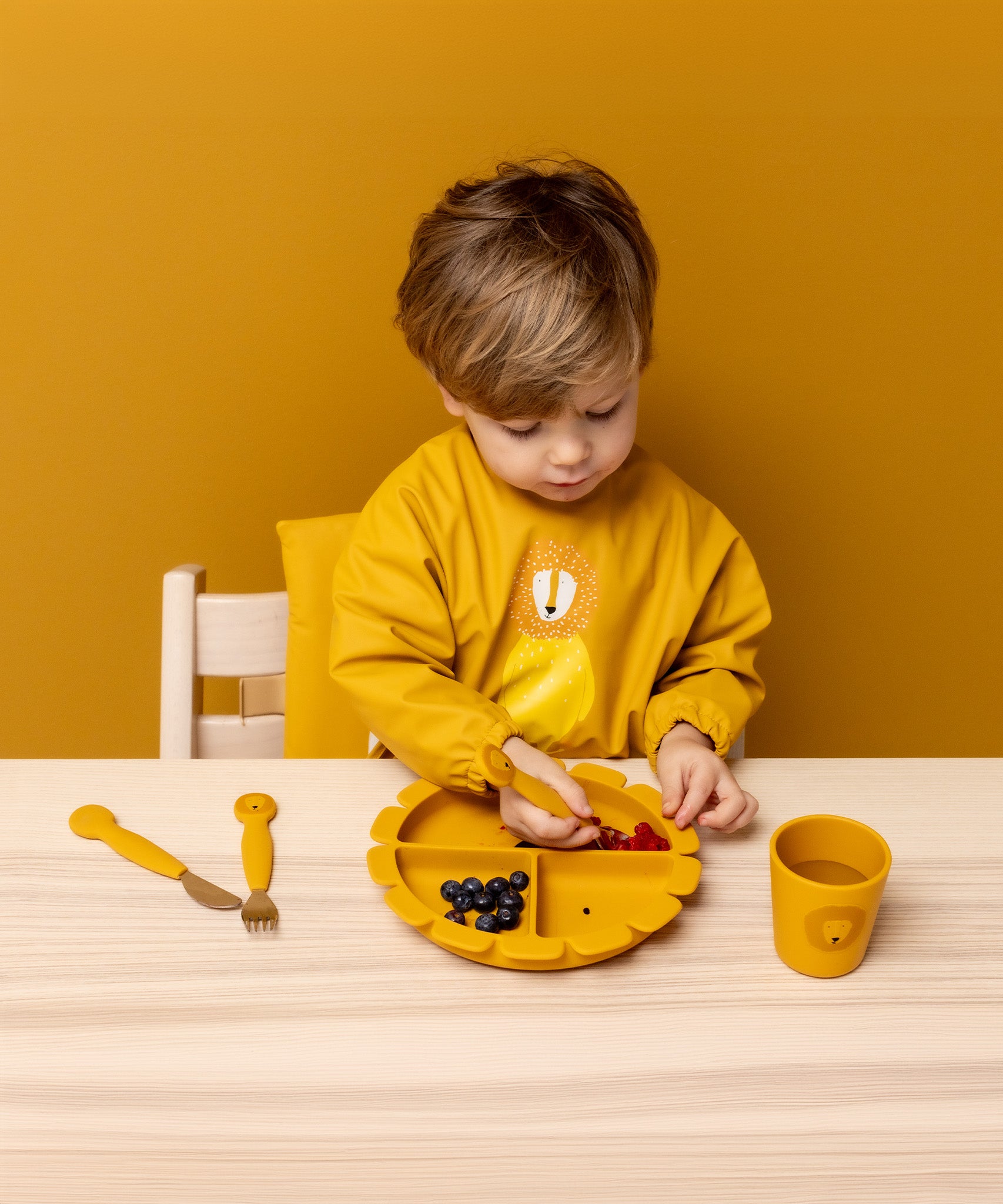 Child wearing Trixie Animal Friends Waterproof Long Sleeve Bib, yellow, Mr Lion, eating from a matching plate and cutlery set. 