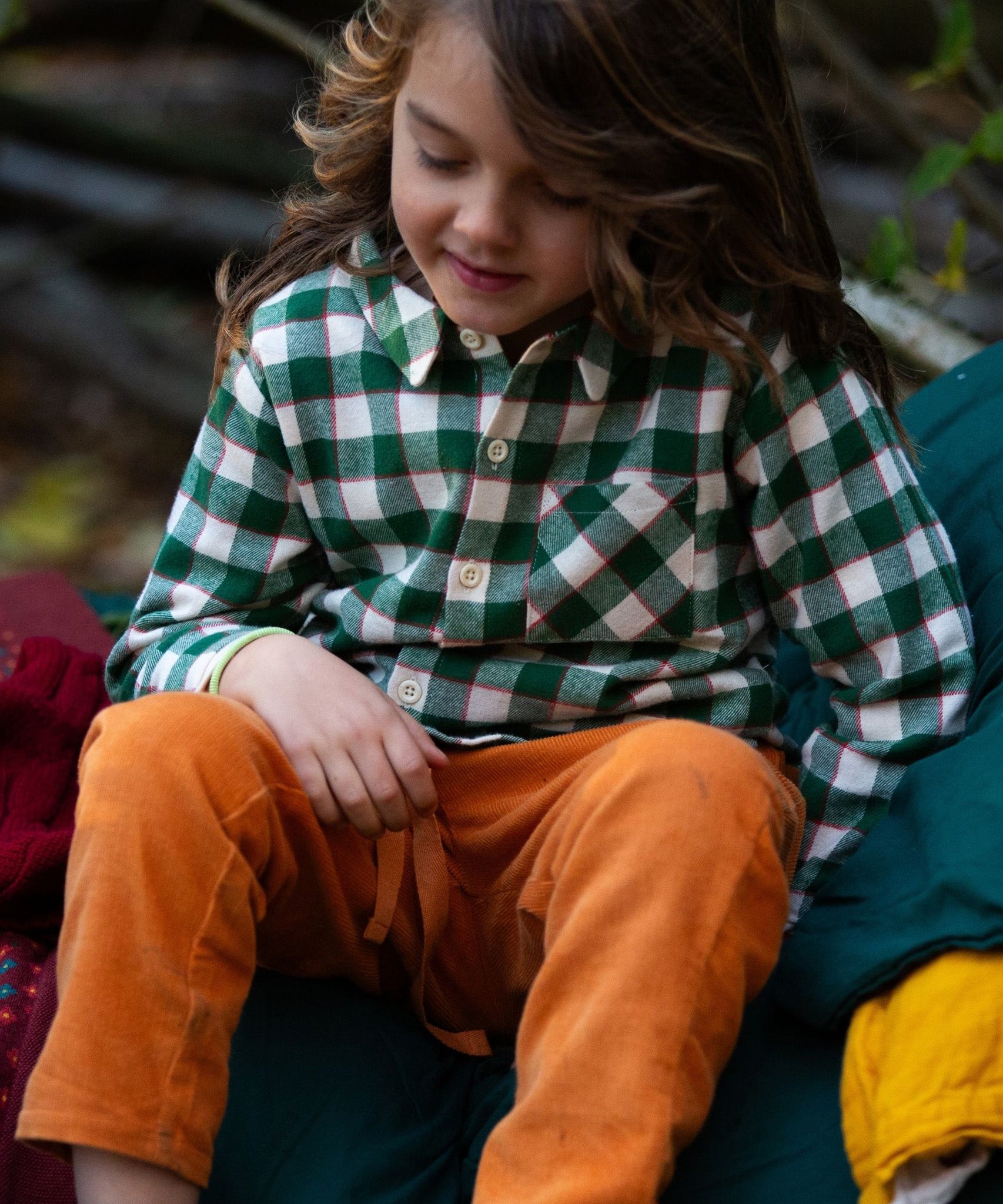 A child sat down, wearing the Little Green Radicals Check Long Sleeve Shirt - Fern Green with yellow trousers