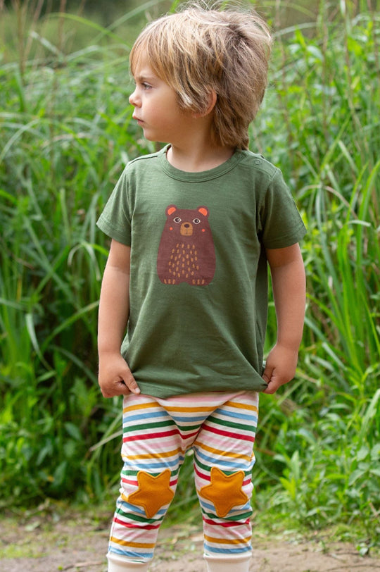 Young child stood outdoors wearing Organic and Fairtrade clothing by ' Little Green Radicals' - A green tshirt with a bear motif and stripey joggers with star knee patches. 