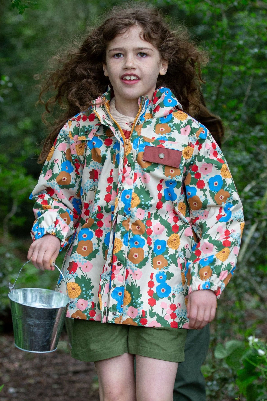 Child exploring a lush green forest wearing a Little Green Radicals waterproof raincoat with a vibrant floral print. Designed for outdoor adventures, this eco-friendly coat is fluorocarbon-free, windproof, and features taped seams for superior waterproof protection. With a breathable and durable design, it keeps little explorers dry and comfortable in all weather. The child carries a small metal bucket, embracing nature play. Available at Babipur, this sustainable raincoat combines style, function, and ethi