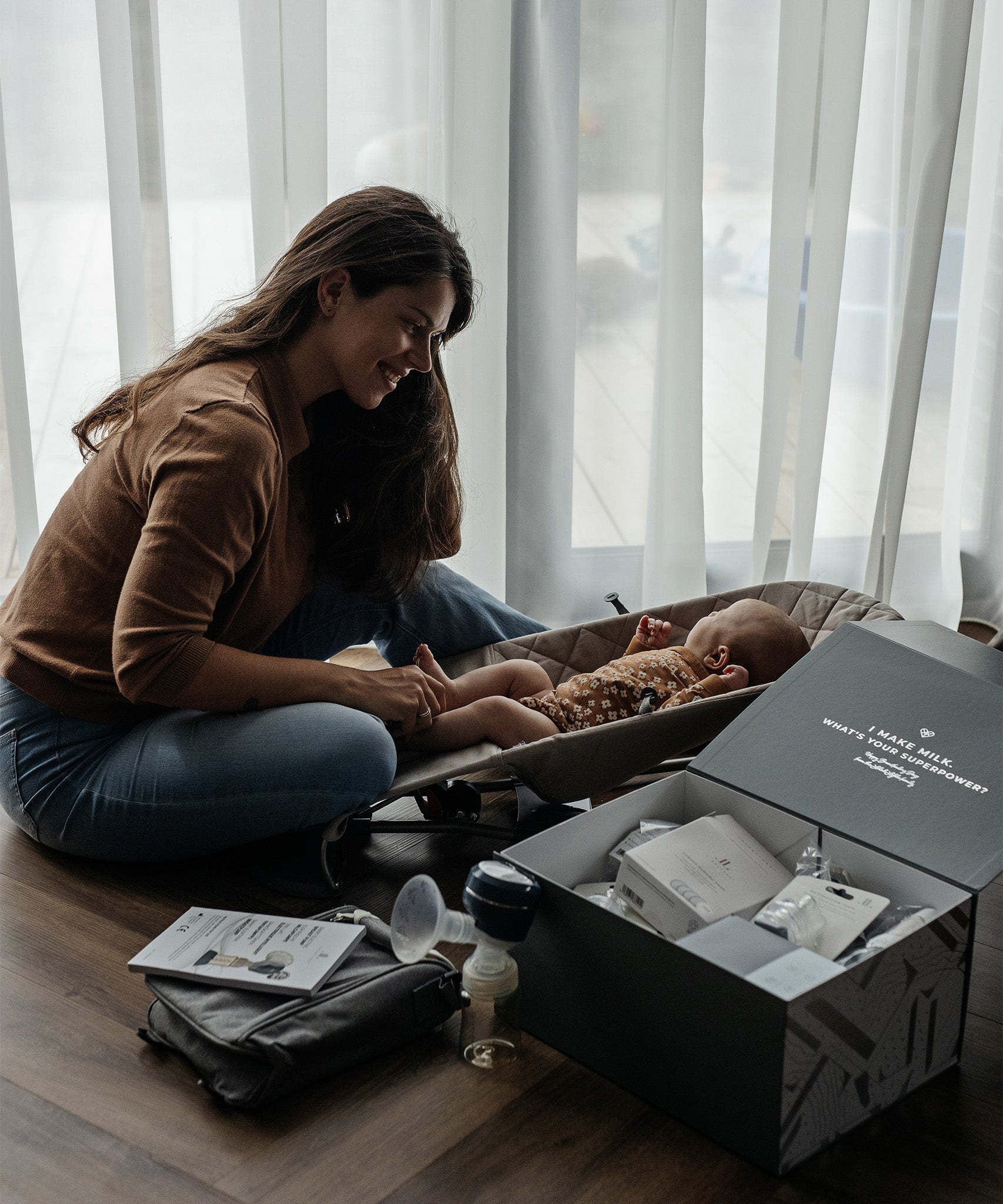 A person sits in front of their child which is in a baby boucer chair, with the Lola & Lykke Breastfeeding Starter Kit next to them