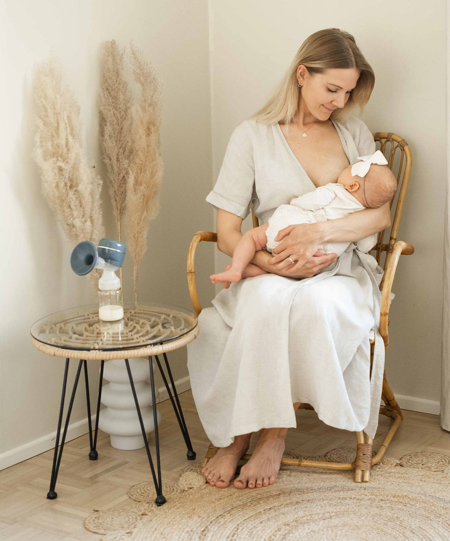 An adult breastfeeding a baby, with the Lola&Lykke Smart Electric Breast Pump on a table next to them.