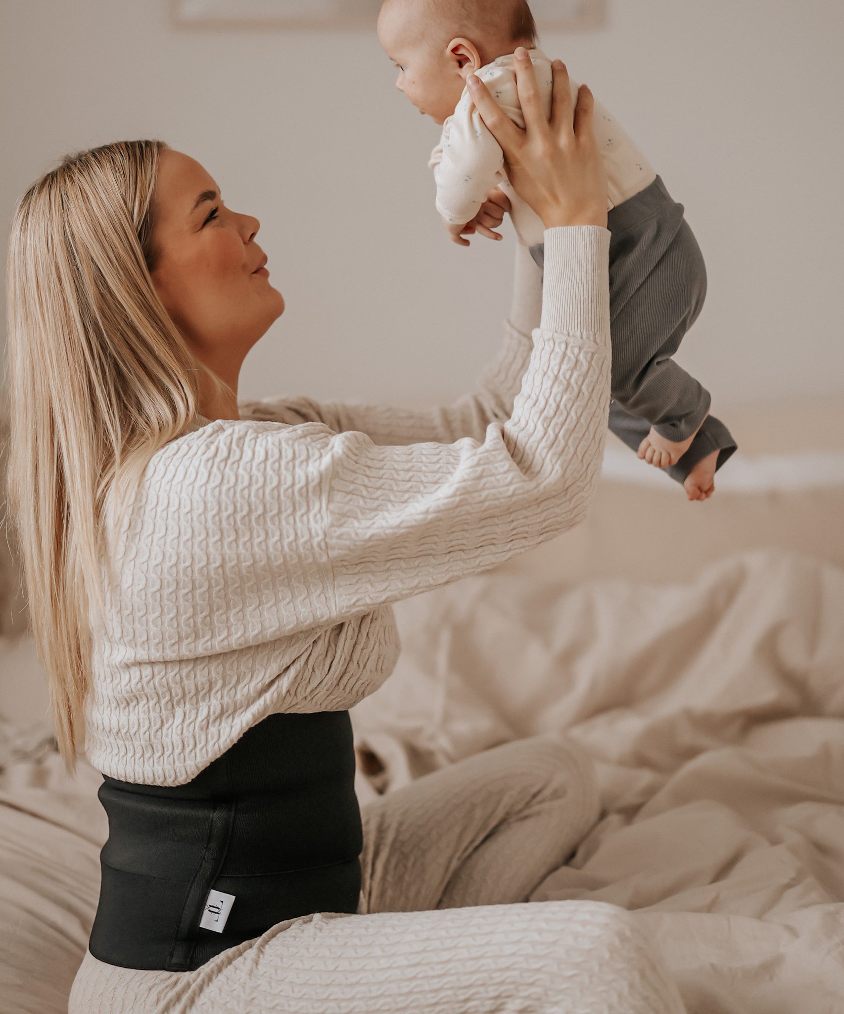 A person holding a baby in the air, wearing the Lola & Lykke Core Restore Postpartum Belly Band