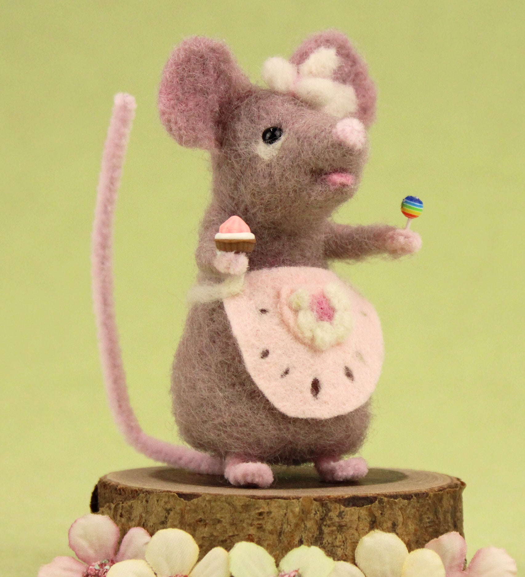 The Makerss Candy Mouse holding a cupcake and a lollypop, and wearing a pink apron on a green background