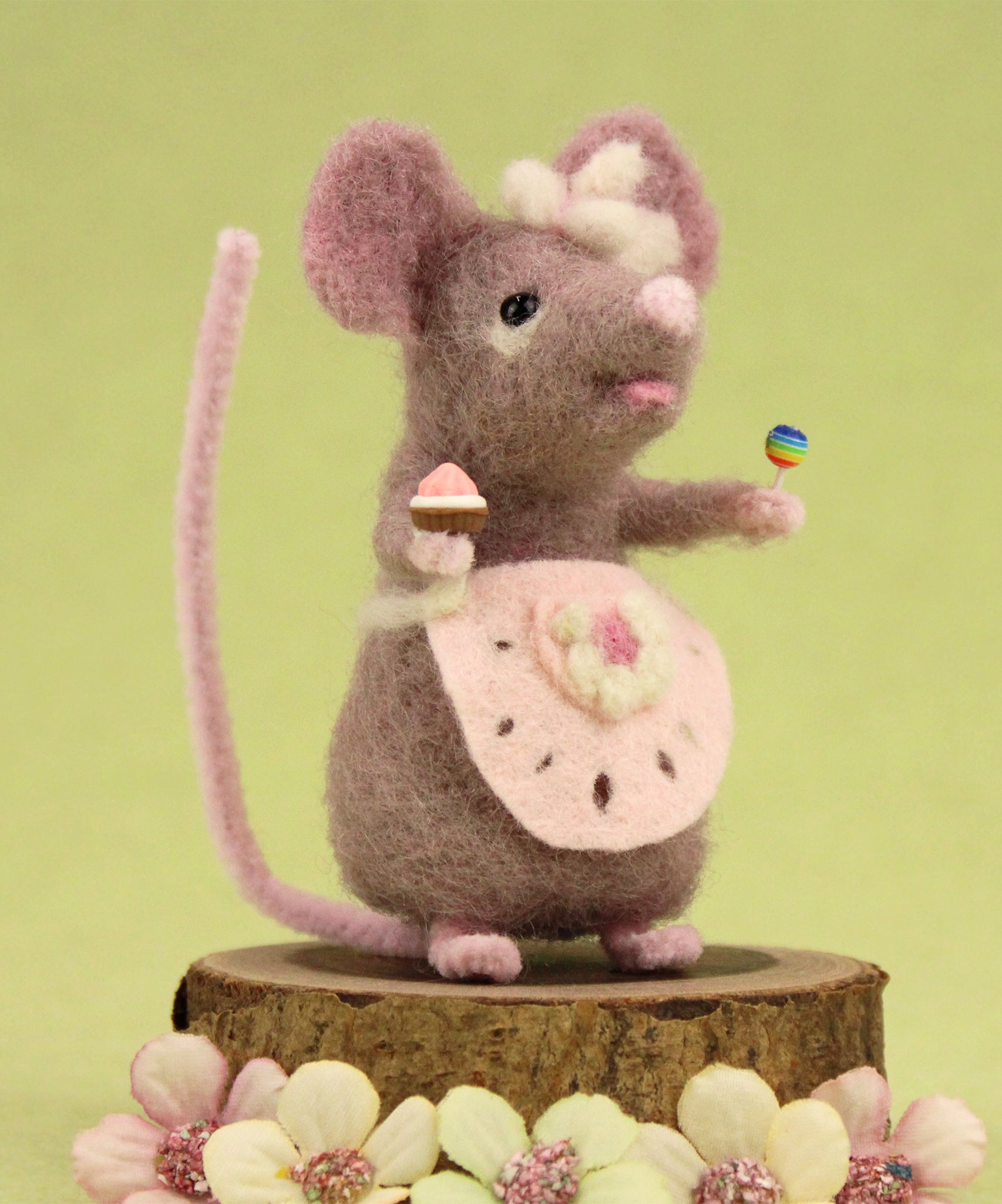 The Makerss Candy Mouse holding a cupcake and a lollypop, and wearing a pink apron on a green background