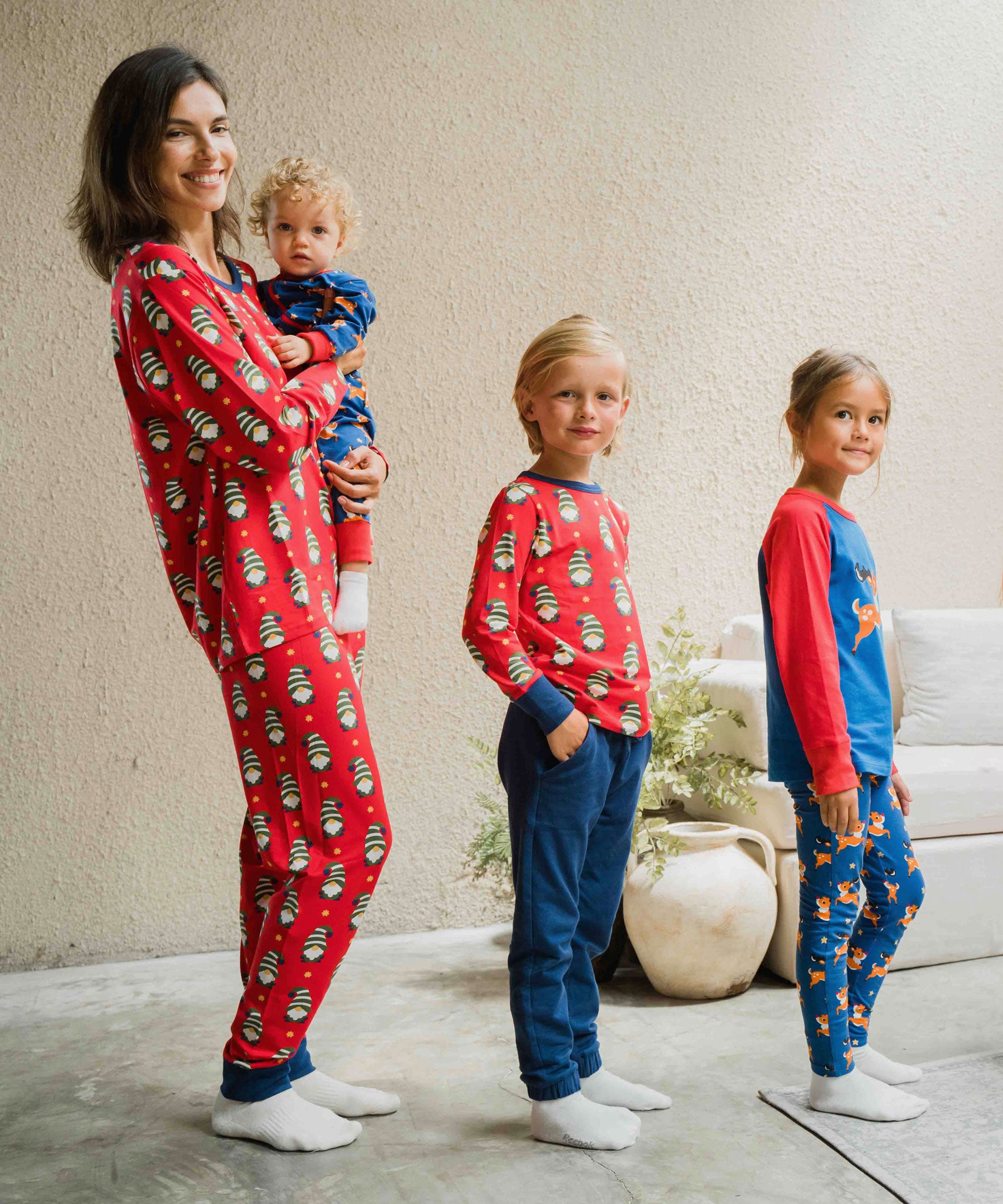 An adult and three children all wearing items from the Maxcomorra Christmas collection. The adult is wearing the Swedish Santa pyjamas, holding a baby wearing the reindeer print rompersuit. One of the other two children is wearing a Swedish Santa top and the last child is wearing a reindeer raglan top with reindeer print leggings. 