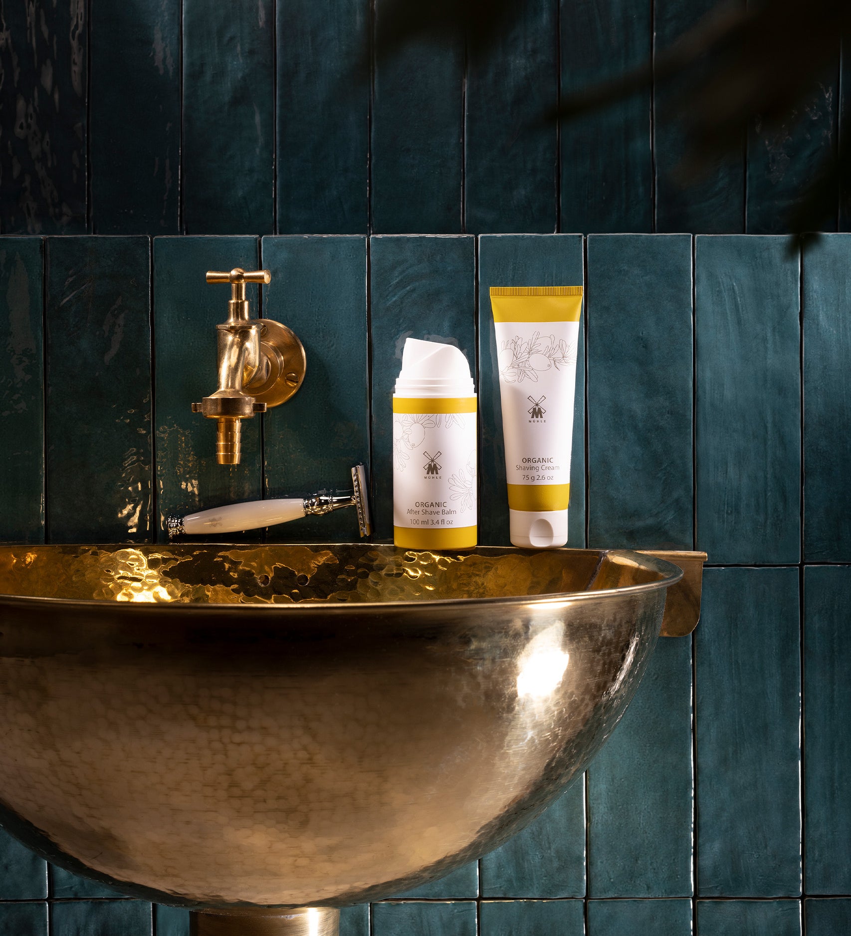 MÜHLE Organic Shaving Cream is stood on a golden sink next to the MÜHLE Organic Shaving Foam, on a dark blue tiled background