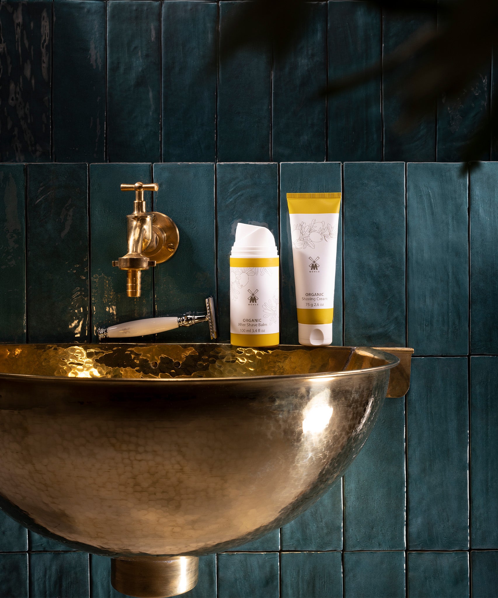 MÜHLE Organic Shaving Cream is stood on a golden sink next to the MÜHLE Organic Shaving Foam, on a dark blue tiled background