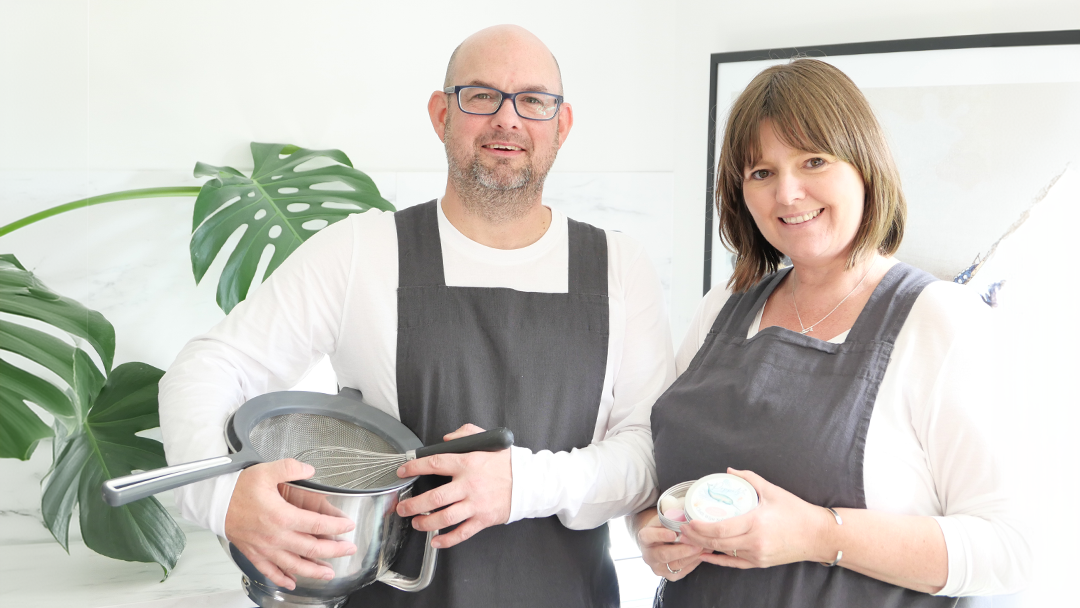 Poppet's creators husband and wife team pose together.  Mr Poppets has a bowl and whisk in his hands and Mrs Poppets has a finished product in tin 