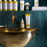 MÜHLE Organic Shaving Cream is stood on a golden sink, next to the MÜHLE Organic Serum, with other MÜHLE products above on a shelf. The background is of dark blue tiles