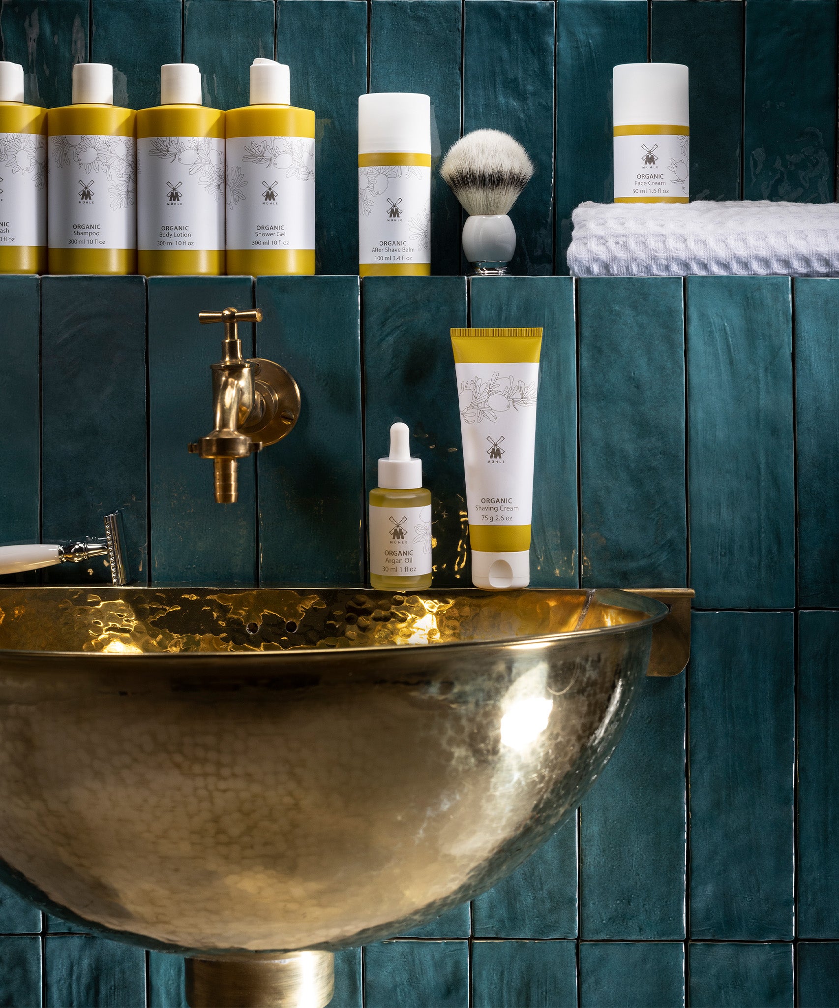 MÜHLE Organic Shaving Cream is stood on a golden sink, next to the MÜHLE Organic Serum, with other MÜHLE products above on a shelf. The background is of dark blue tiles