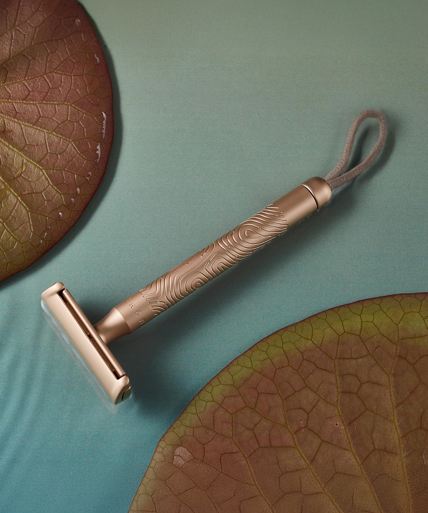 MÜHLE Companion Razor - Rose Gold, is sat on a blue table that looks like water, with two water lilies either side 