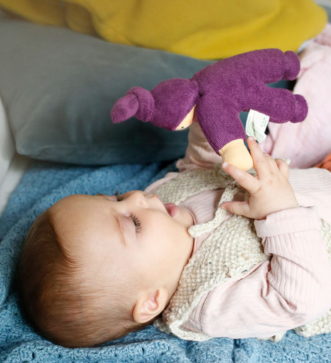 A baby playing with a purple Nanchen Waldorf Pimpel doll, looking at the face of the doll in delight