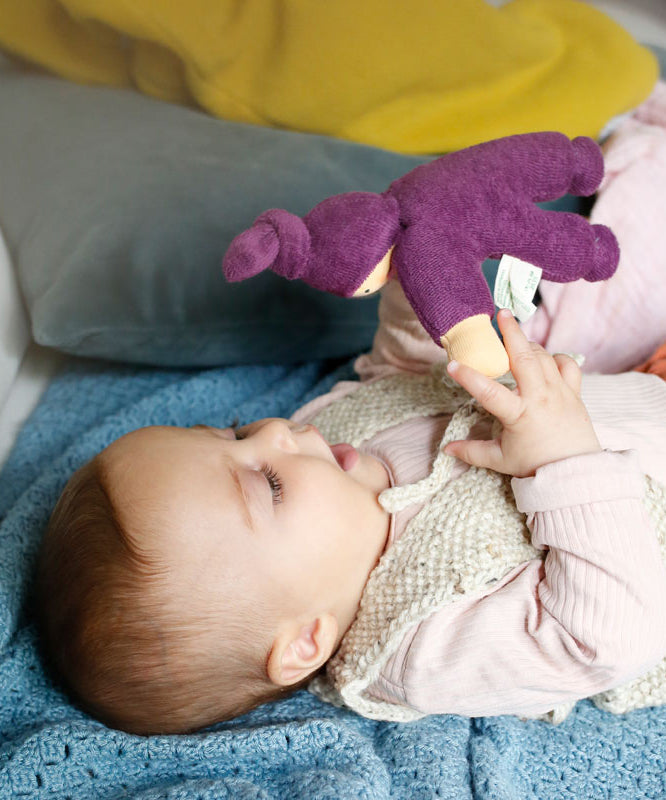 A baby playing with a purple Nanchen Waldorf Pimpel doll, looking at the face of the doll in delight