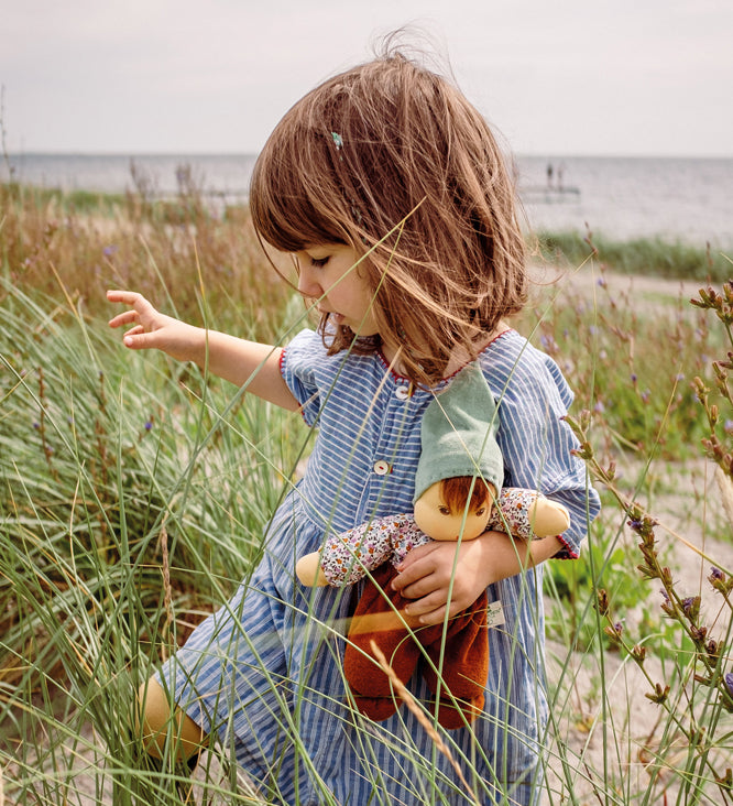 A child holding a Nanchen Forest Child Waldorf Doll in their arms, whilst walking along a grassy beach area