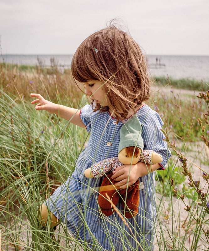 A child holding a Nanchen Forest Child Waldorf Doll in their arms, whilst walking along a grassy beach area