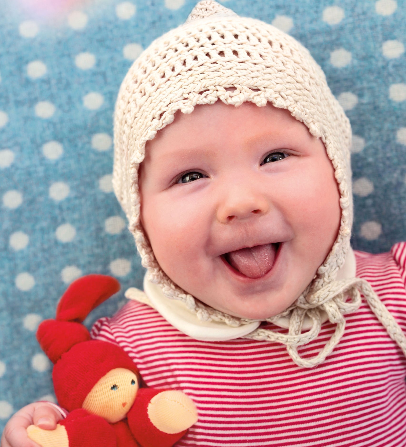 A child is happily lying down on a light blue and white spot blanket, with a big smile on their face, and is wearing a crochet cream bonnet and red stripe top. The child is holding The Nanchen Waldorf Pimpel Doll in red