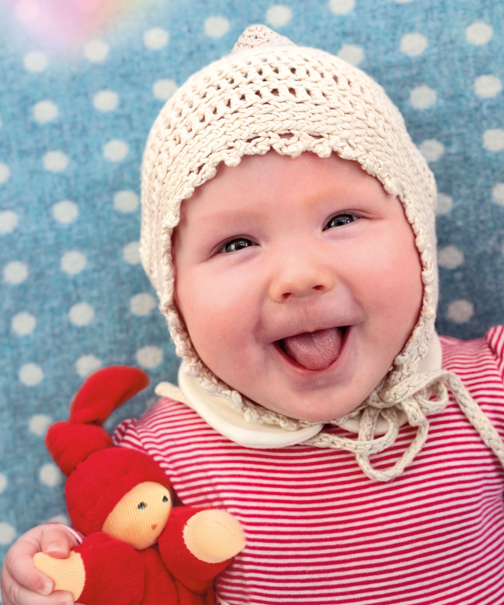 A child is happily lying down on a light blue and white spot blanket, with a big smile on their face, and is wearing a crochet cream bonnet and red stripe top. The child is holding The Nanchen Waldorf Pimpel Doll in red