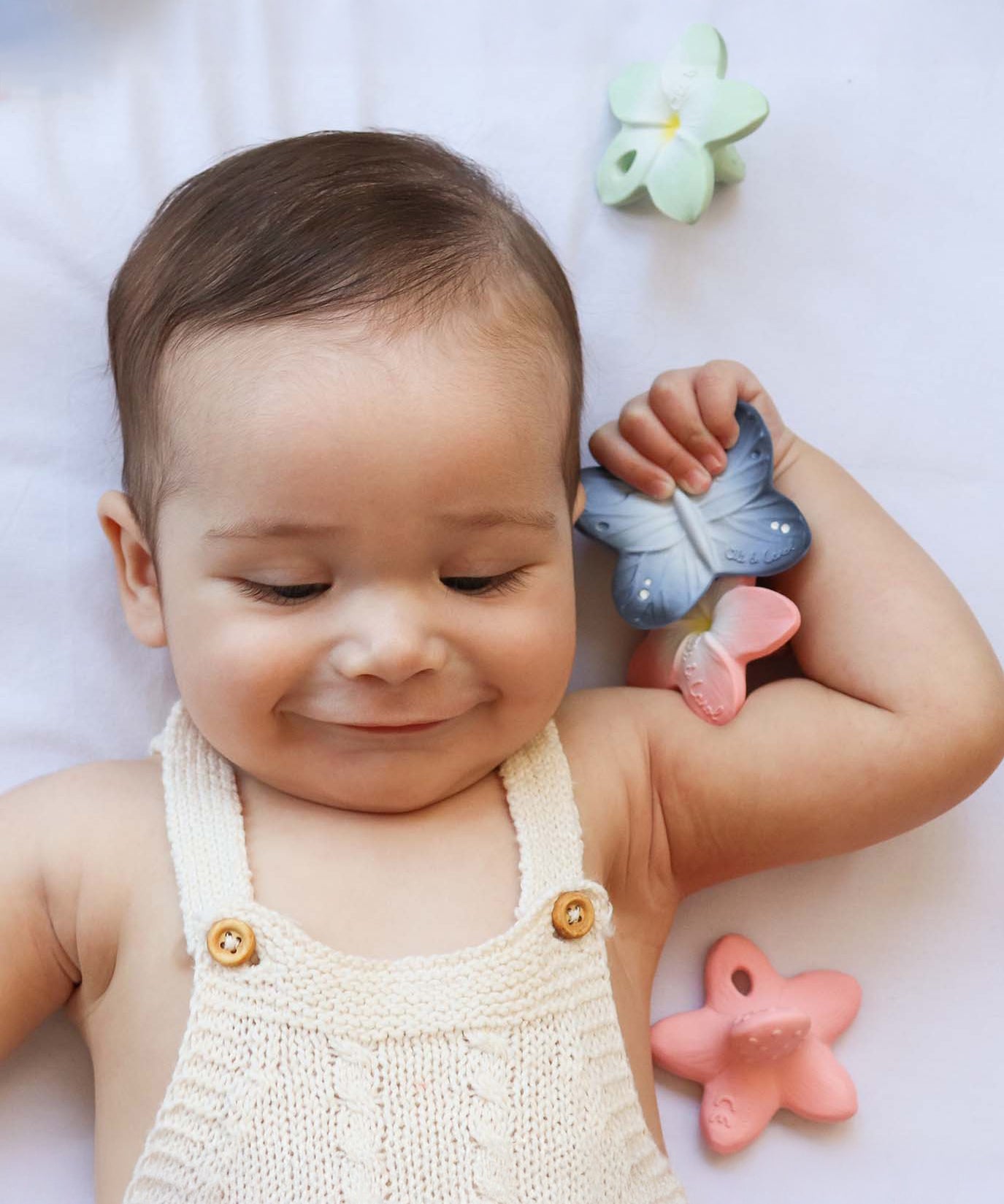 A child smiling and holding the Oli & Carol Natural Rubber Mini Teether - Blues The Butterfly in their hand. Around the child are other Oli & Carol natural rubber teethers