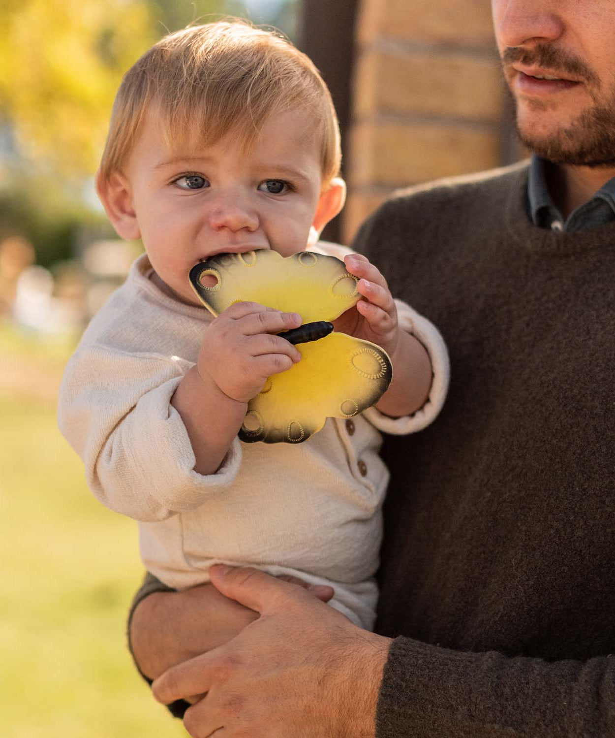 A child being held by an adult, chewing on the yellow Oli & Carol Natural Rubber Teether - Katia The Butterfly