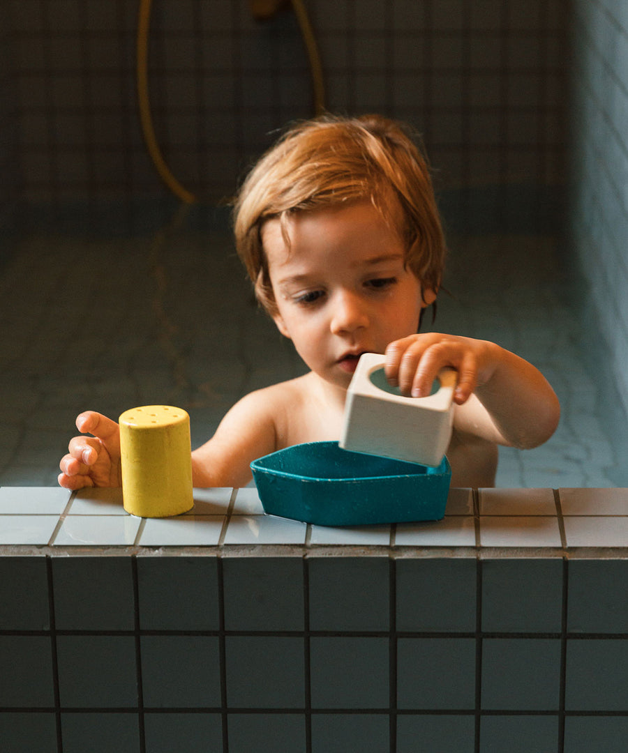 A child in a bath, playing with the blue Oli & Carol Boat Bath Toy, made from 100% Upcycled Natural Rubber. The child is playing the parts of the boat together