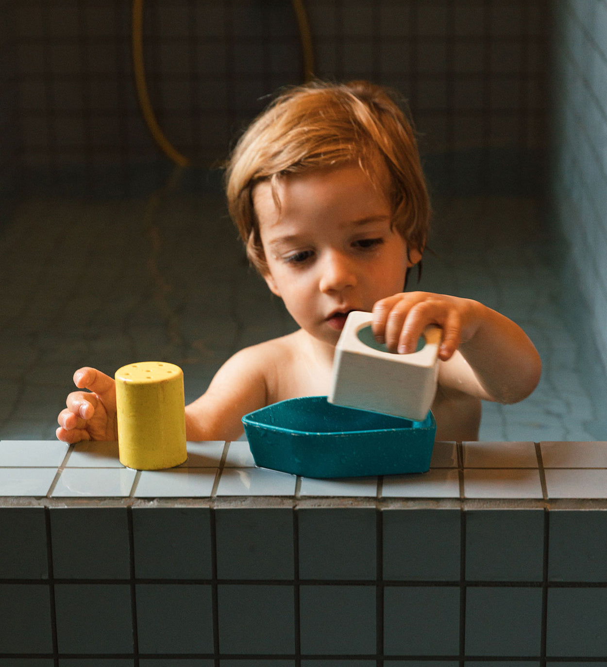 A child in a bath, playing with the blue Oli & Carol Boat Bath Toy, made from 100% Upcycled Natural Rubber. The child is playing the parts of the boat together