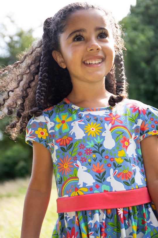 A young girl with curly hair smiles outdoors, wearing a lilac Frugi Issey Party Skater Dress Bunny Bounce - made from GOTS organic cotton  with a bunny, rainbow, and floral print, featuring cap sleeves and a pink waistband.