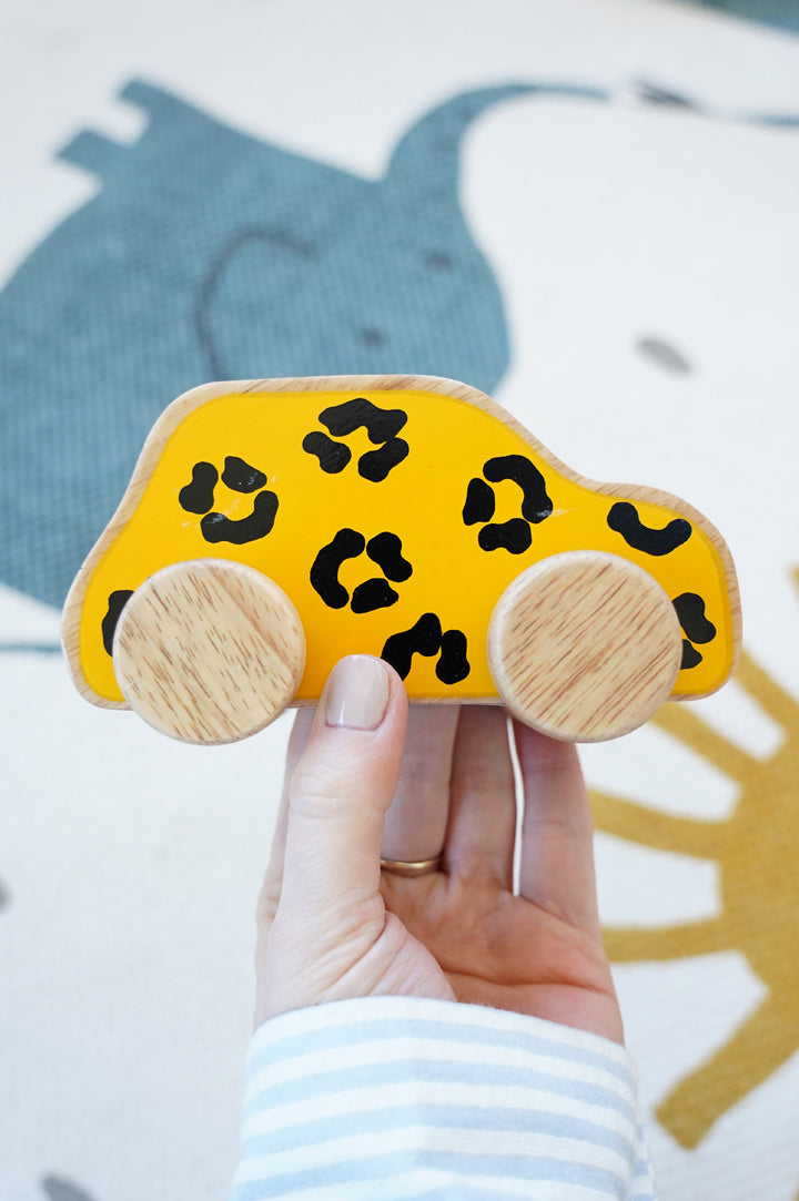 Lanka Kade push along rubber wood car held in a hand over a cream rug. The body of the car is a bold yellow and black leopard print, the wheels are left natural, showing the grain of the wood.