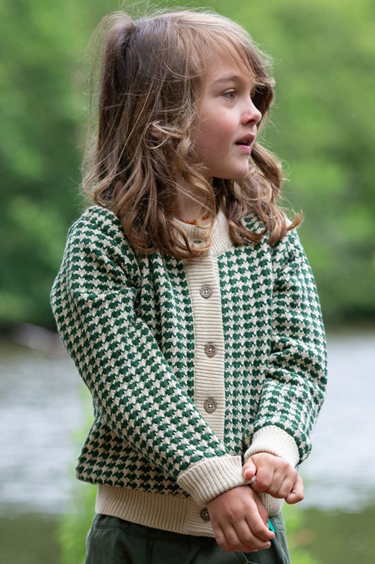 A child wearing the Little Green Radicals Olive Striped Cardigan—made from Fairtrade and organic True Knit cotton—stands in a forest