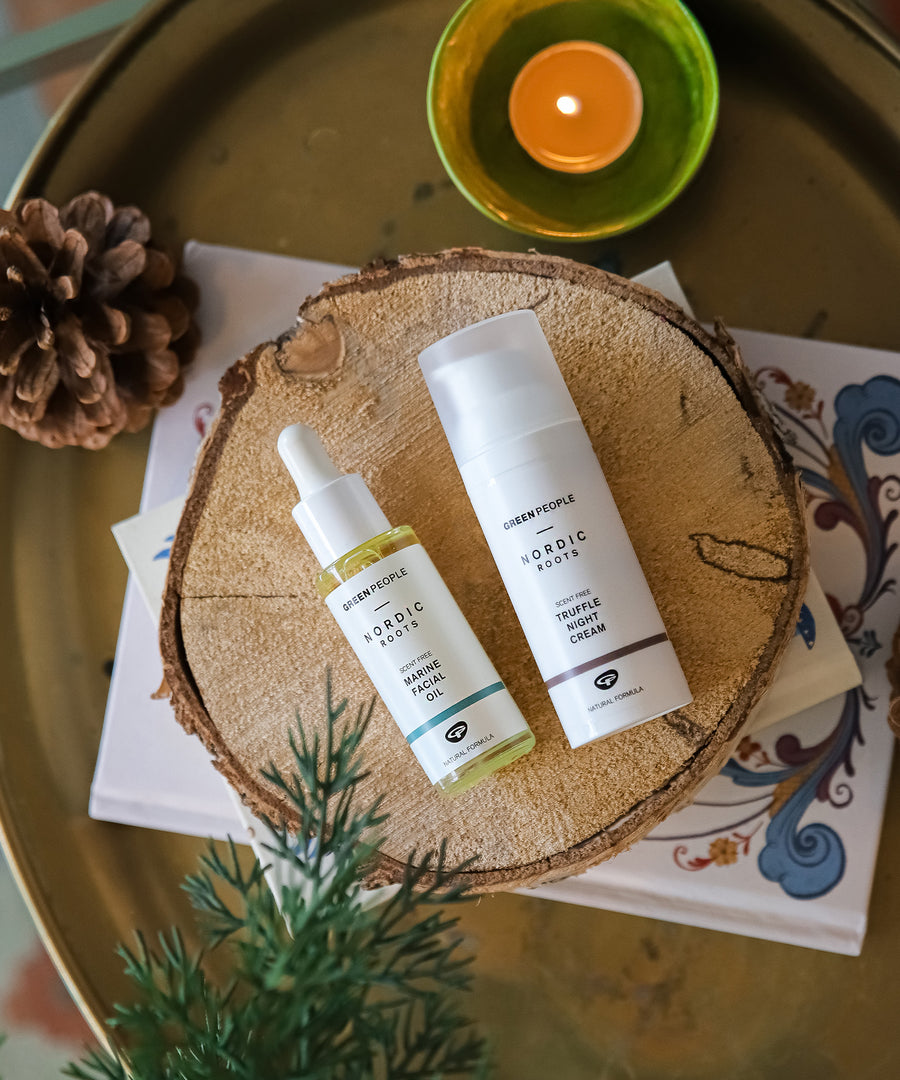 The Green People Nordic Roots Marine Facial Oil and Truffle Night Cream are laid on a wooden disk with a candle, pine cone and spruce tree branch surrounding them