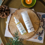 The Green People Nordic Roots Marine Facial Oil and Truffle Night Cream are laid on a wooden disk with a candle, pine cone and spruce tree branch surrounding them