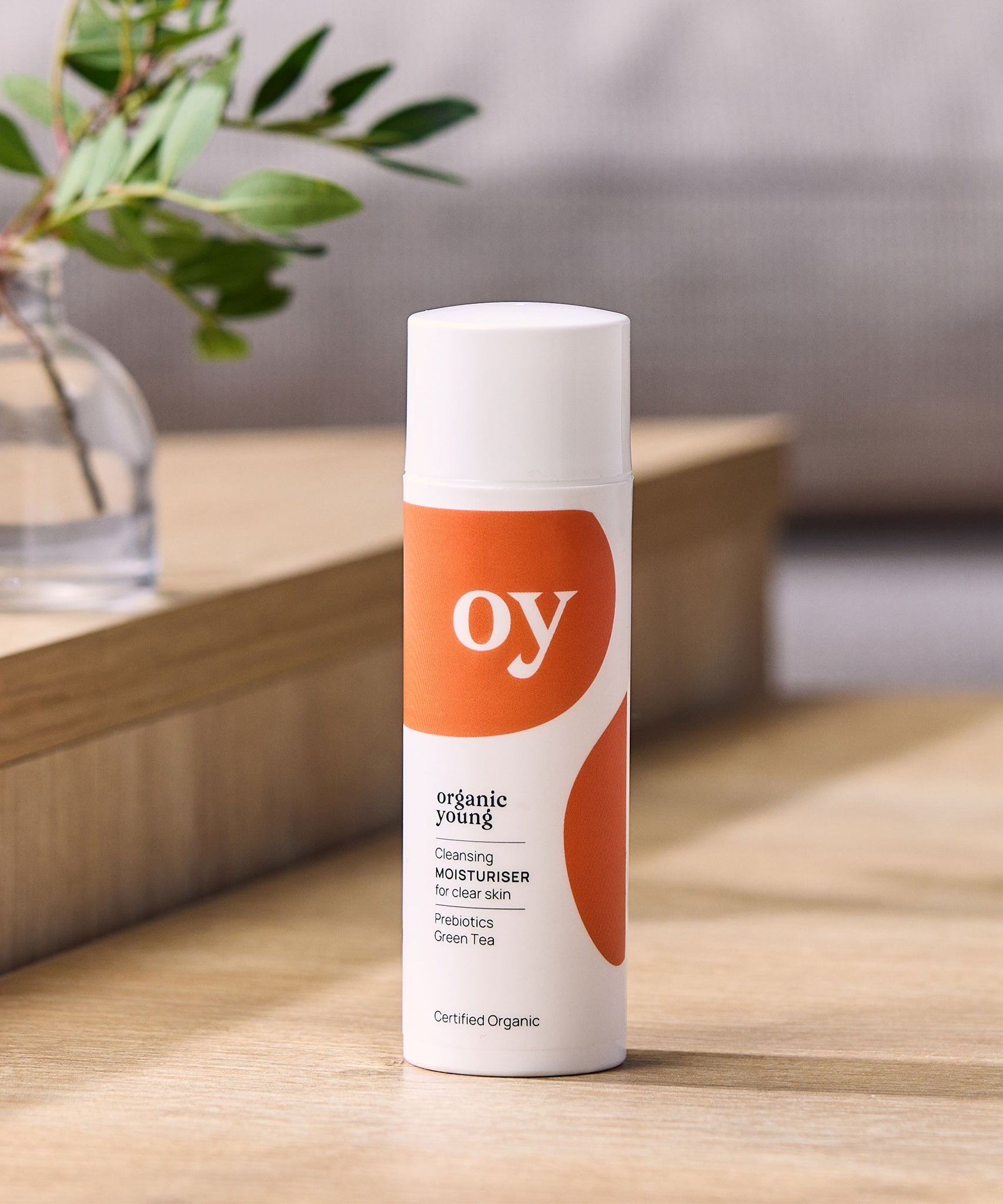 The Oy organic cleaning moisturiser for clear skin with prebiotics and green tea, is stood on a brown table with a plant in the background