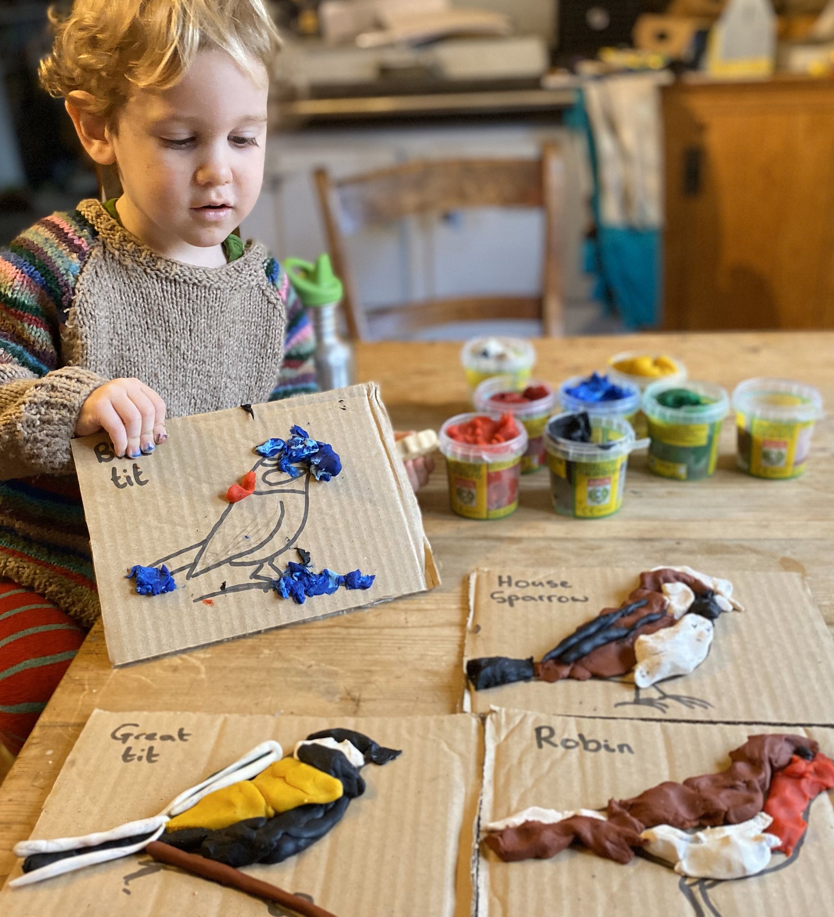 Child showing art he has created using Okonorm play dough. The child has recreated different birds on cardboard by using the different coloured play doughs. The child is sat on a chair at the kitchen table. He has one piece of art in his hands, the other three are on the table.