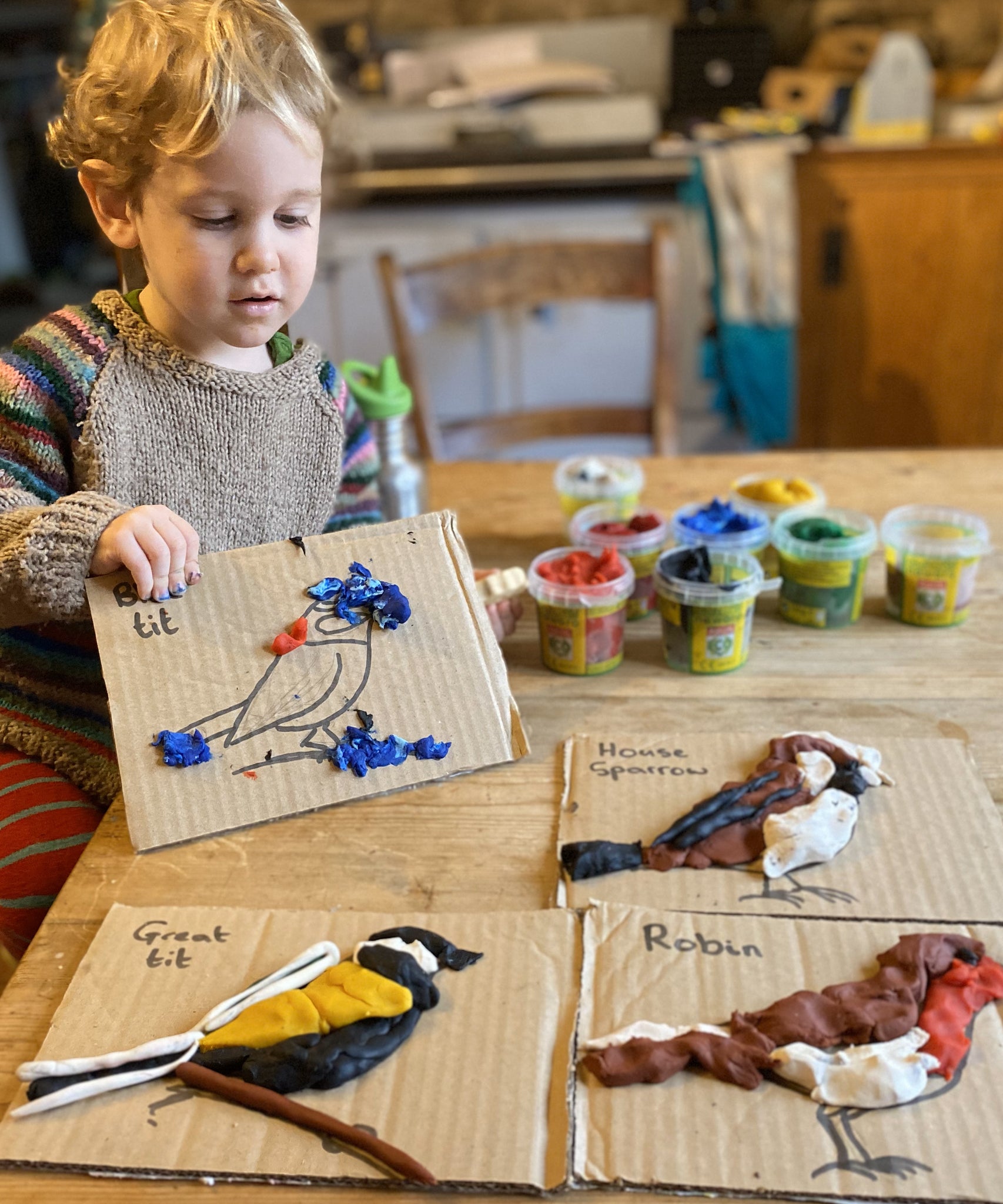 Child showing art he has created using Okonorm play dough. The child has recreated different birds on cardboard by using the different coloured play doughs. The child is sat on a chair at the kitchen table. He has one piece of art in his hands, the other three are on the table.