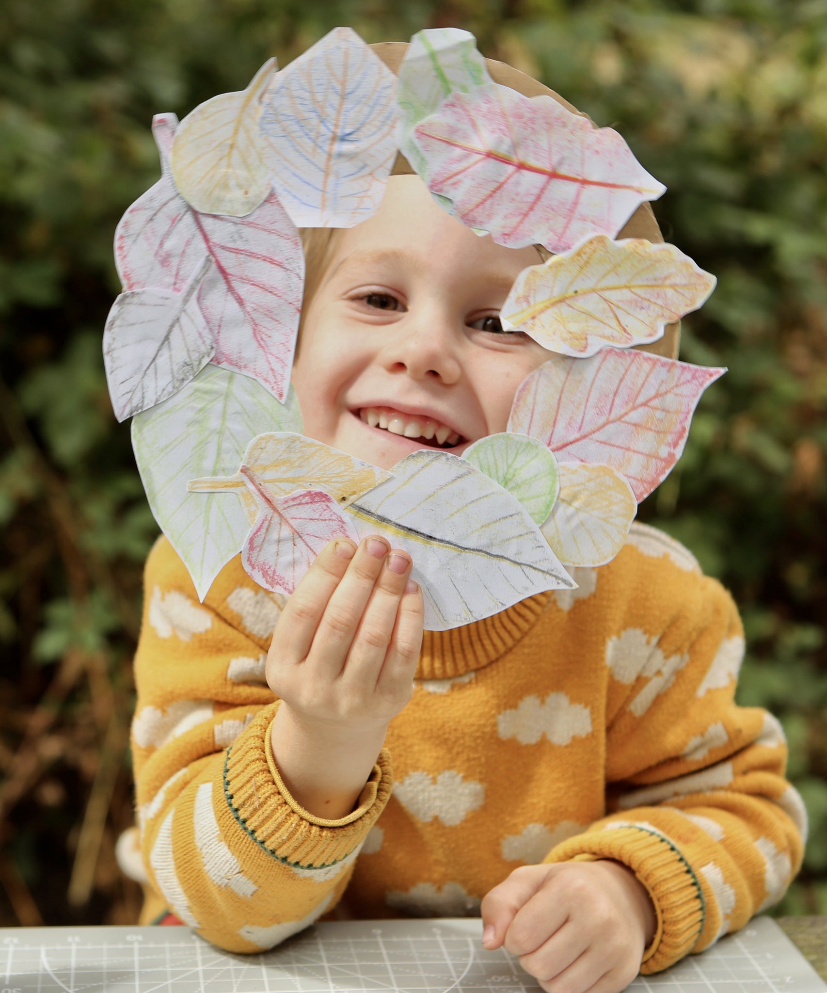 Child holding a paper leaf wreath he has made with leaf rubbings, having used Okonorm wax blocks. The child is holding the wreath in front of his face, you can see the child's face in the hole of the wreath. The child is wearing a yellow knitted jumper with a white cloud pattern and is sat outside.