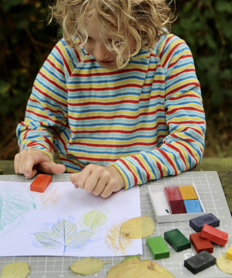 Okonorm wax blocks being used by a child. The child is sat outside in the garden and is creating leaf rubbings with the wax blocks. The child is wearing a striped hoodie and has curly, blonde, above shoulder length hair.