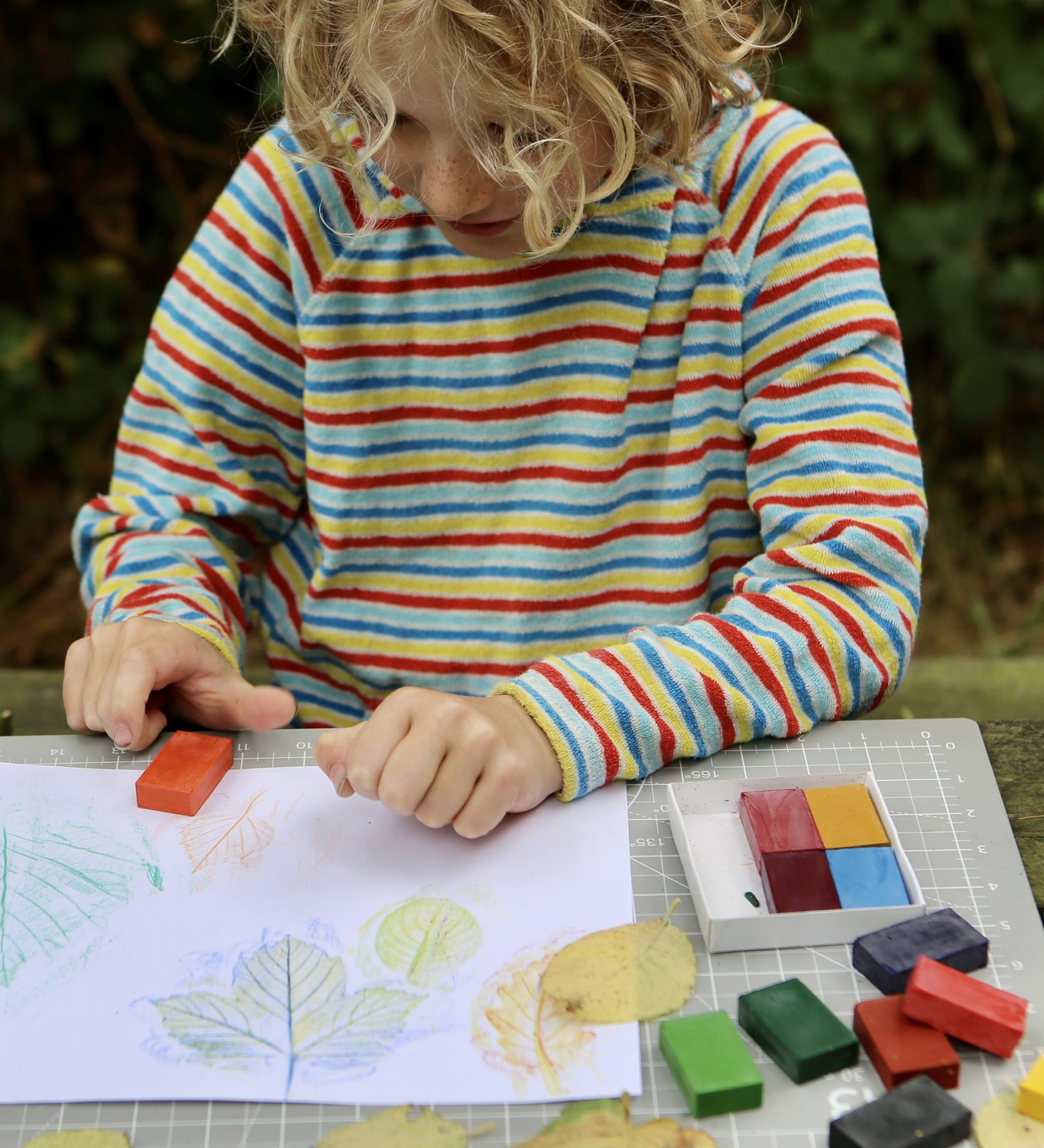 Okonorm wax blocks being used by a child. The child is sat outside in the garden and is creating leaf rubbings with the wax blocks. The child is wearing a striped hoodie and has curly, blonde, above shoulder length hair.