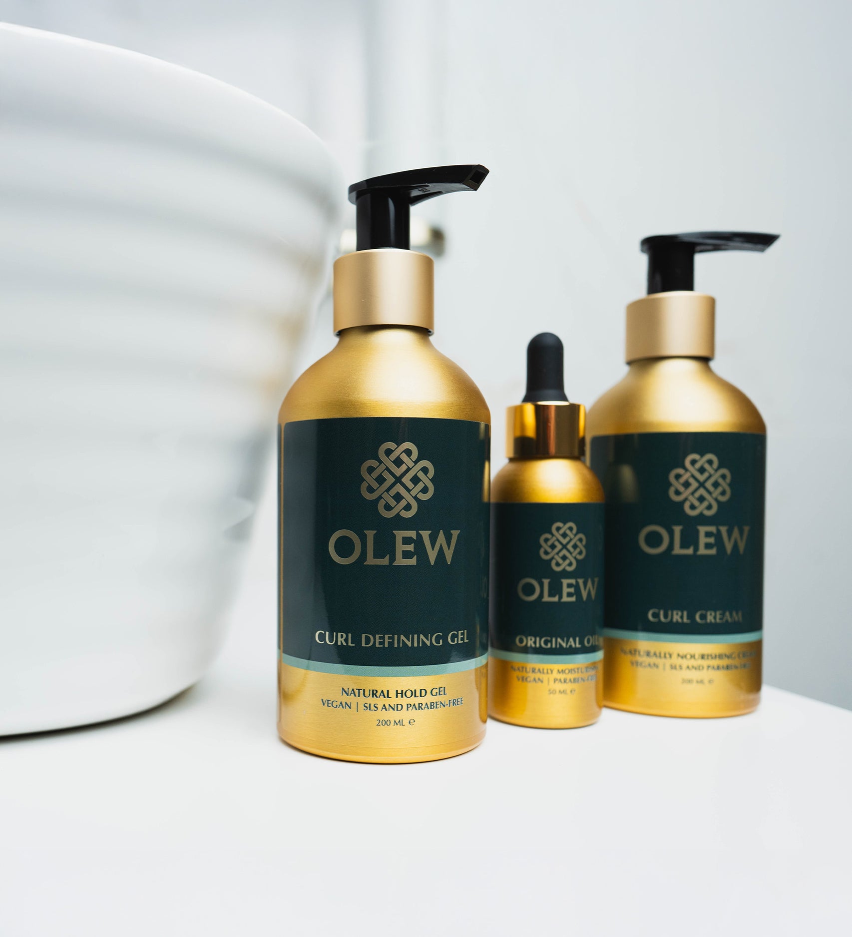 The Olew Curl Defining Gel, Original Oil and Curl Cream in gold and dark green bottles, stood on a white table with a white large bowl next to the bottles.