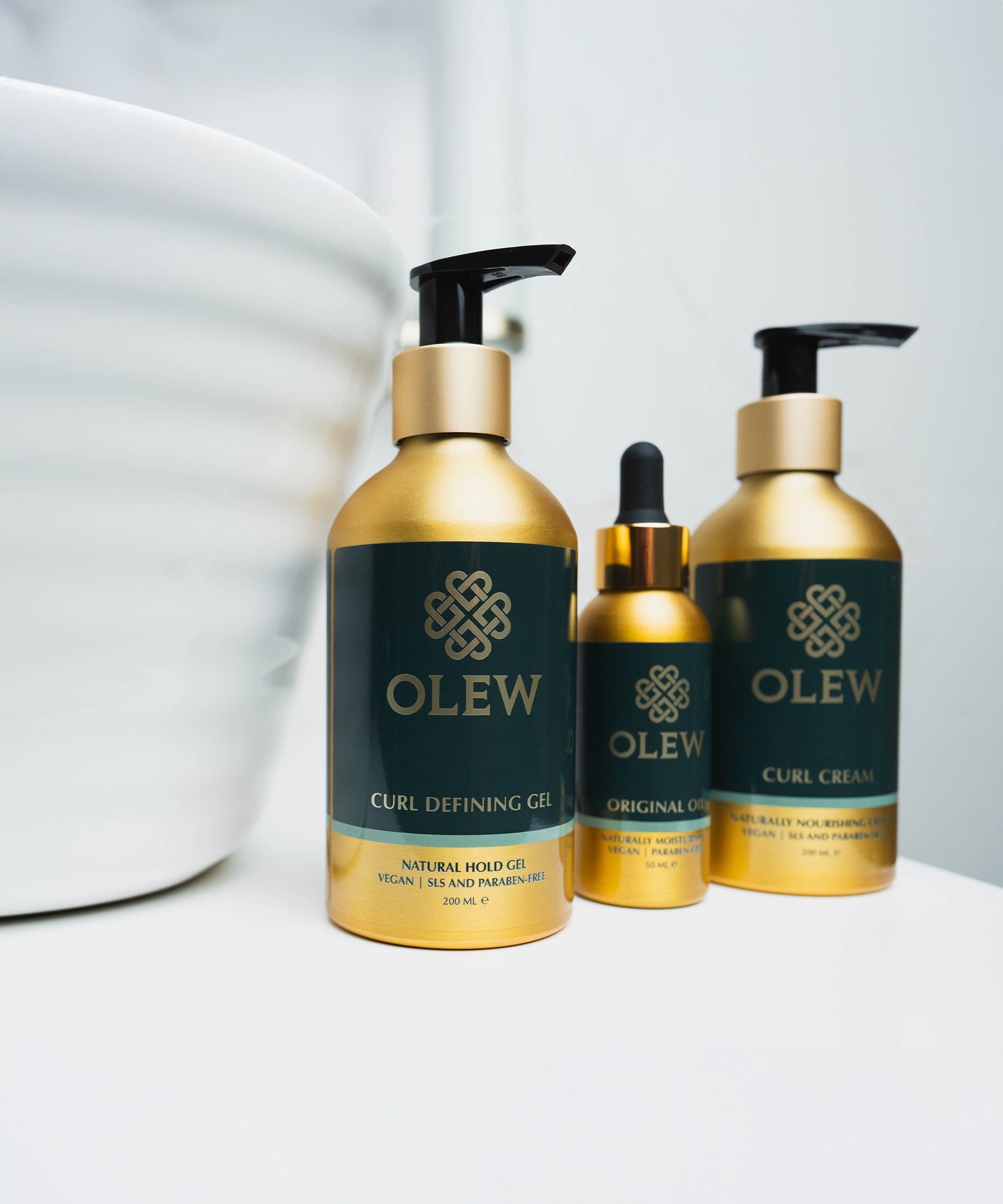 The Olew Curl Defining Gel, Original Oil and Curl Cream in gold and dark green bottles, stood on a white table with a white large bowl next to the bottles.