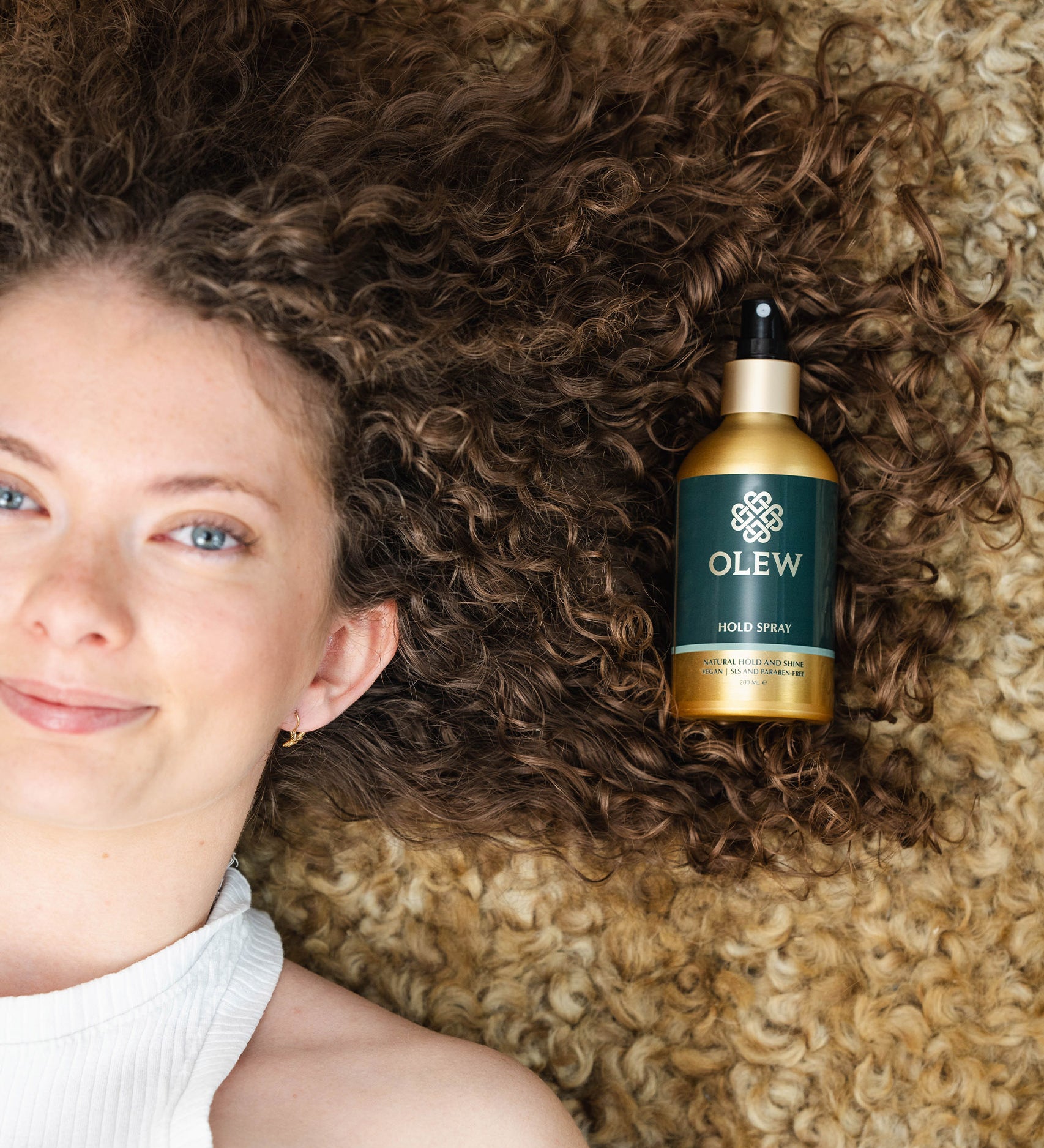 A person with brown curly hair laying down on a light coloured curly rug, with their hair on the rug and the Olew Hold Spray laid on top of their curls.