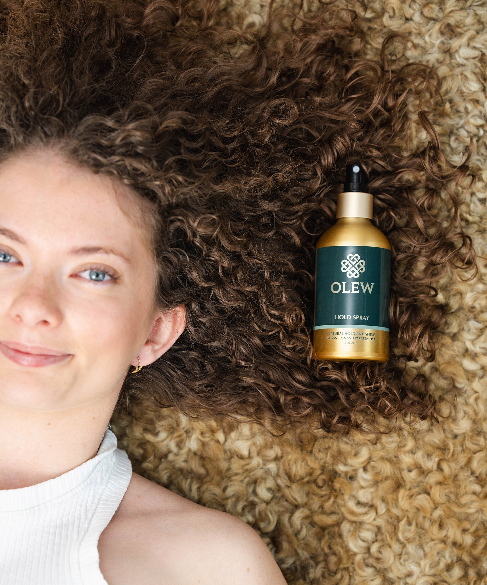 A person with brown curly hair laying down on a light coloured curly rug, with their hair on the rug and the Olew Hold Spray laid on top of their curls.