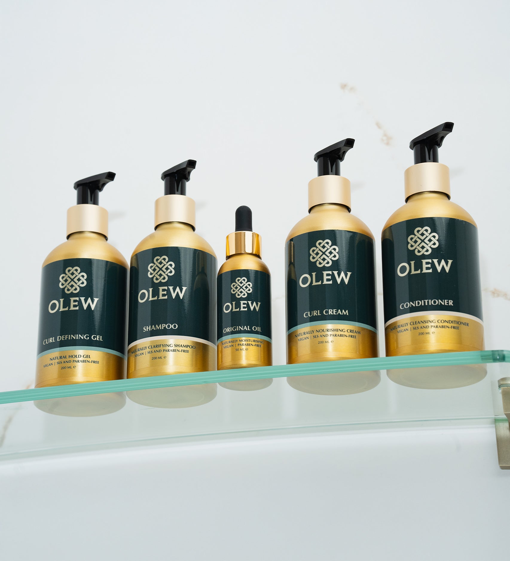 The Olew hair care range together on a glass shelf. The items shown are the Curl Defining Gel, Shampoo, Original Oil, Curl Cream and Conditioner.