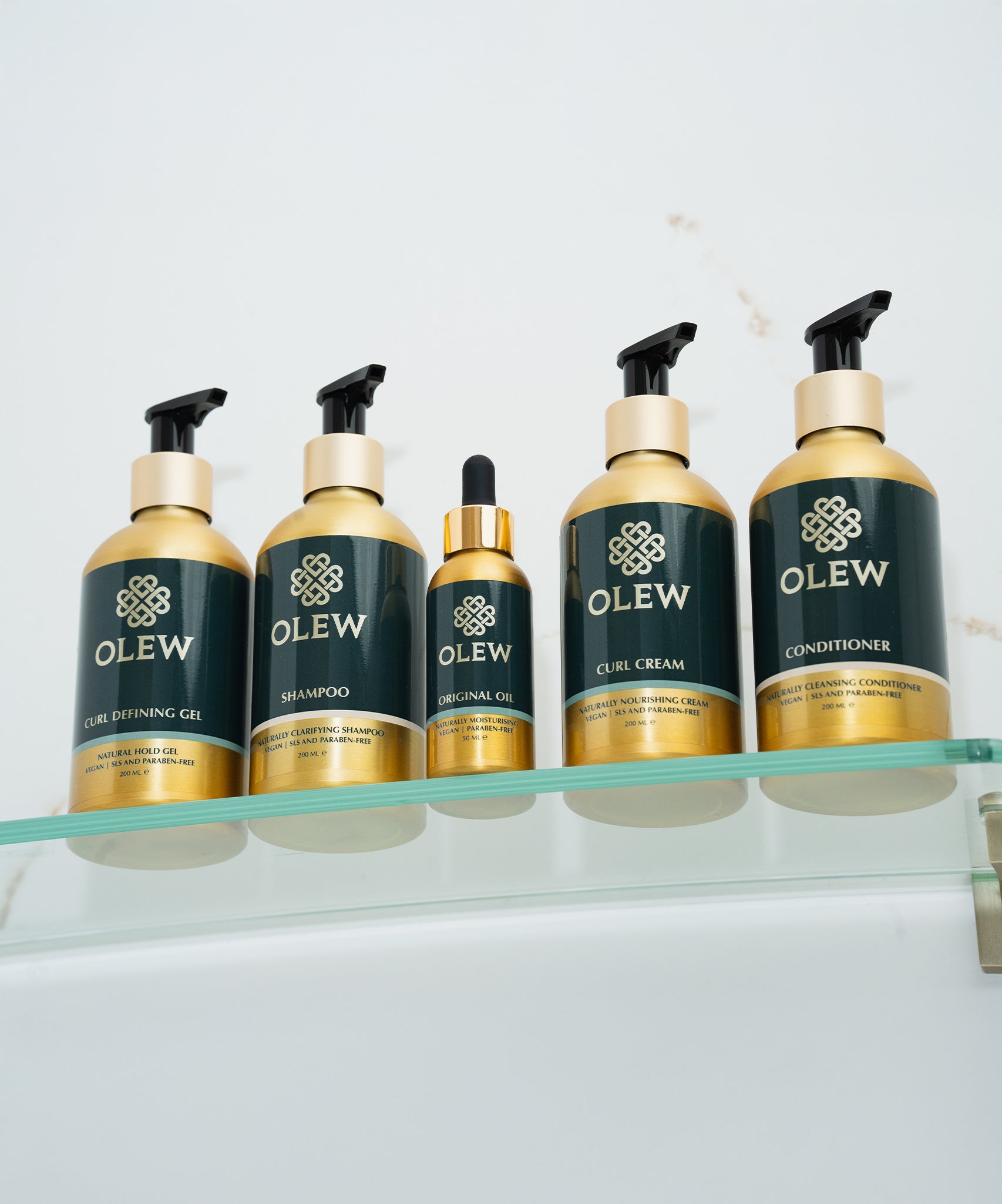 The Olew hair care range together on a glass shelf. The items shown are the Curl Defining Gel, Shampoo, Original Oil, Curl Cream and Conditioner.