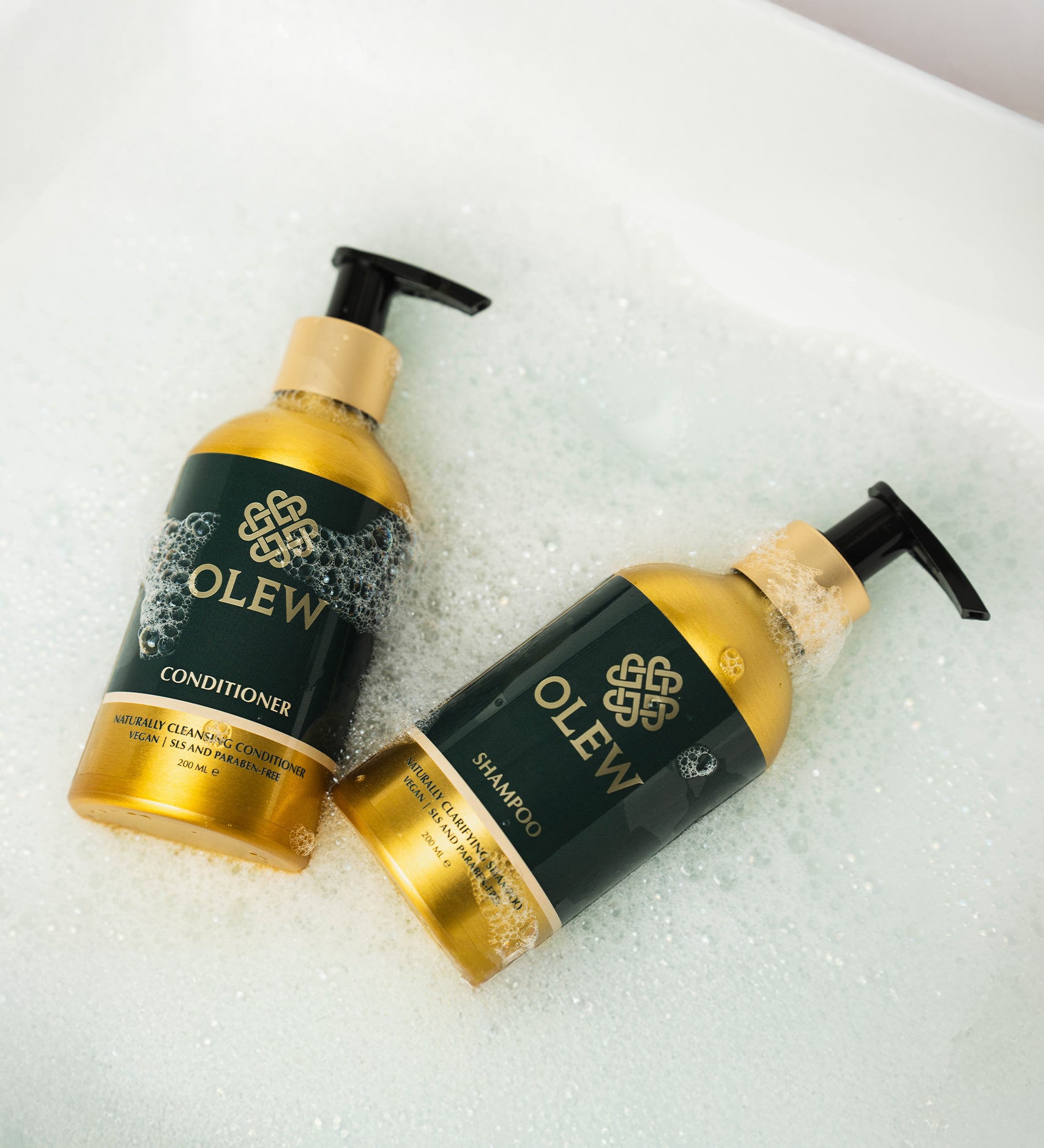 The Olew Shampoo and Conditioner gold and dark green bottles laying in bubbles, to give the feeling of cleanliness