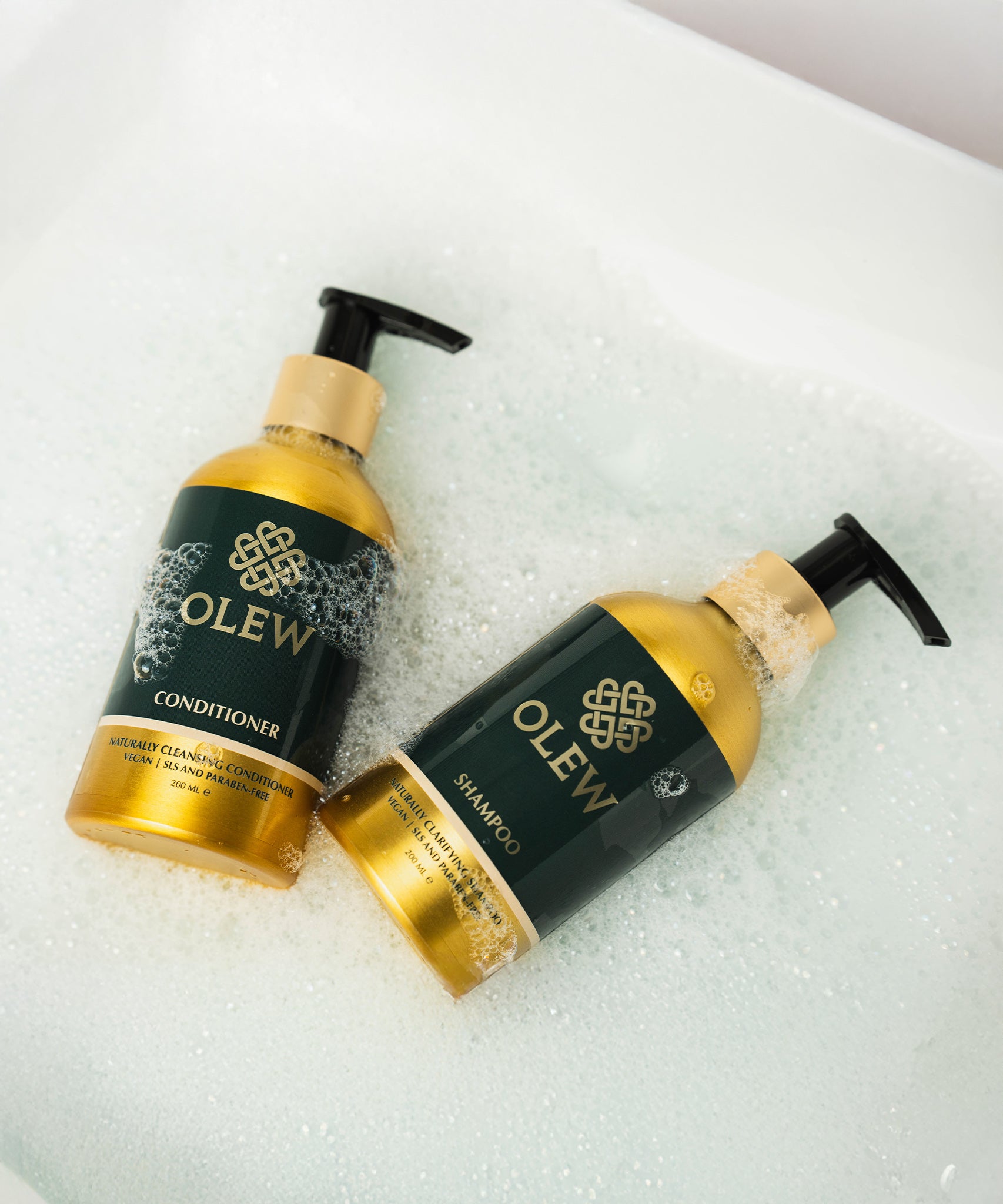 The Olew Shampoo and Conditioner gold and dark green bottles laying in bubbles, to give the feeling of cleanliness