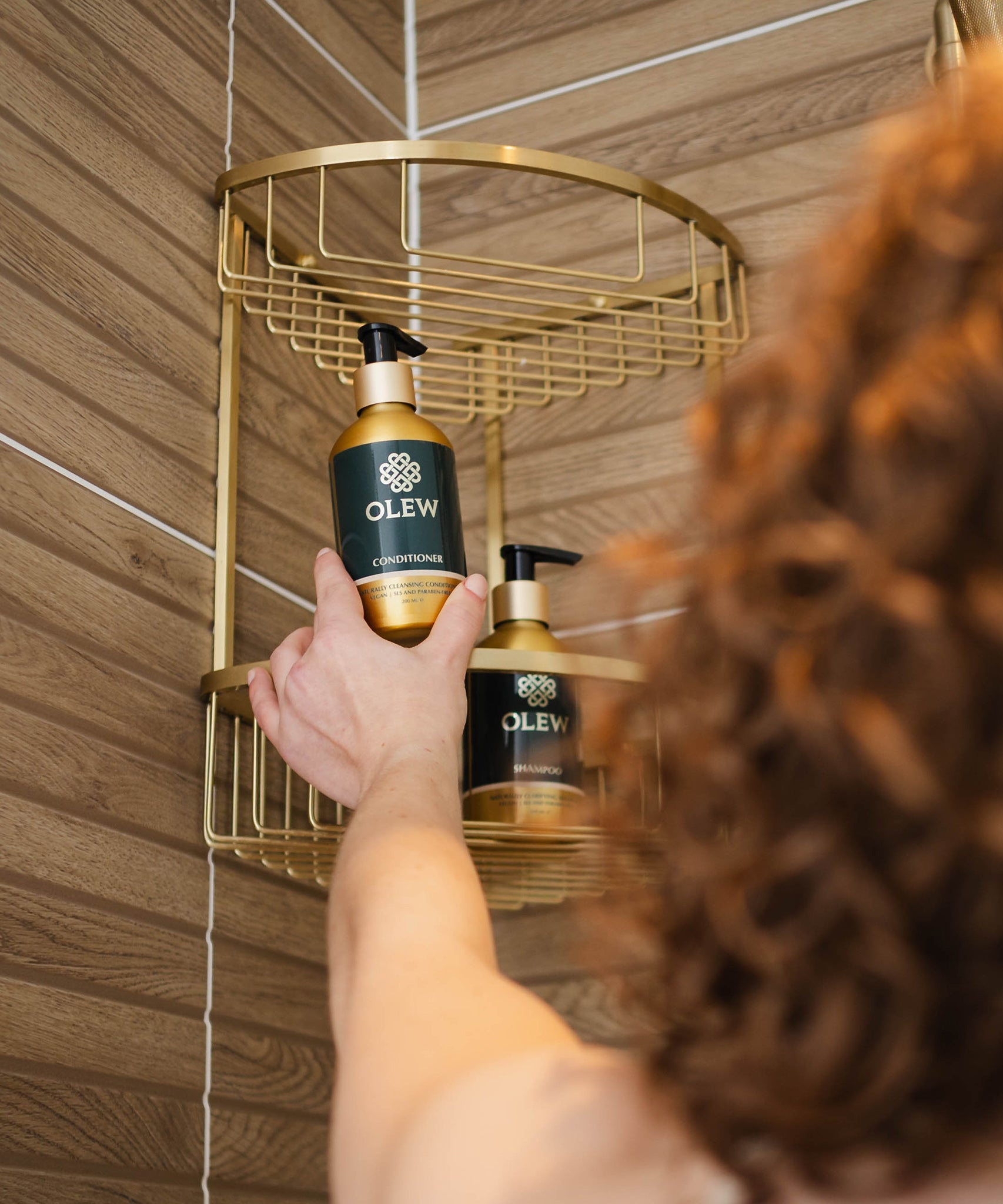 A person reaching for the Olew conditioner and shampoo bottles which are placed in a gold coloured shower caddy