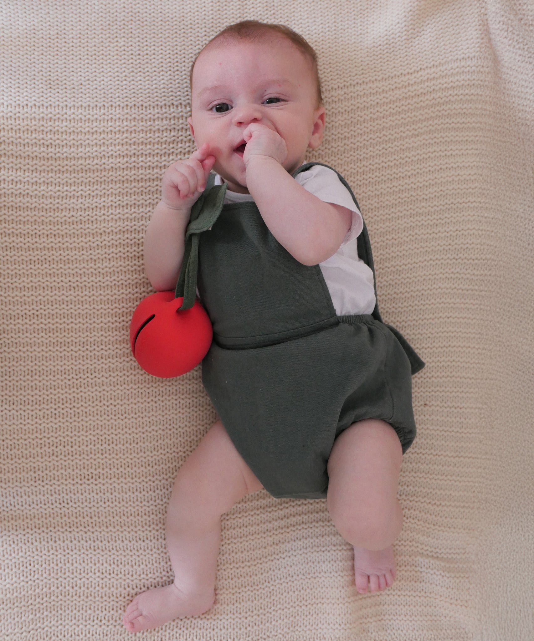 A baby laying down on a cream blanket, playing with the Oli & Carol Pepa Apple Pacifier Holder and Teething Toy